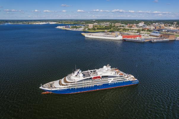 Cruise ship sailing near a harbor under a clear blue sky. The city skyline and docks are visible in the background, evoking a serene atmosphere.