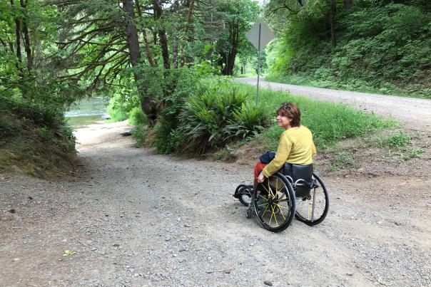 A woman with a wheelchair is on a gravel dirt road leading downhill toward a river.