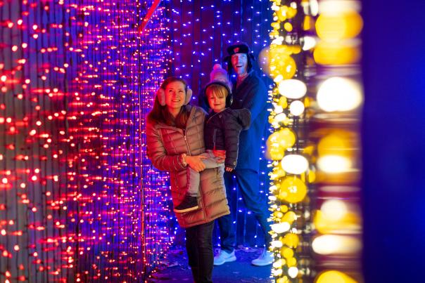 A mom and dad each holding one of their kids as they walk through a tunnel of holiday lights at Henry Vilas Zoo.