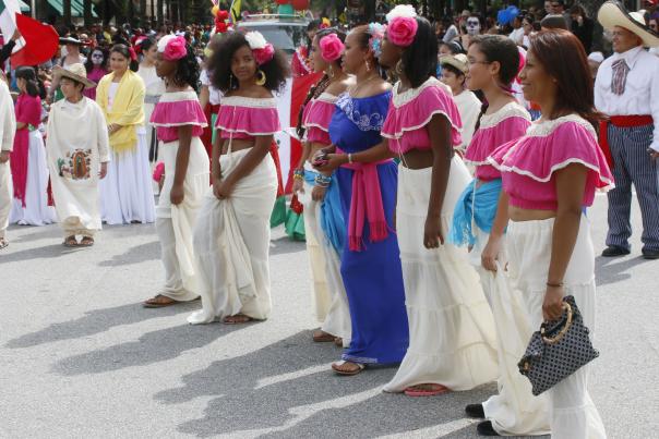 A group of young women in traditional folkloric dresses and bright pink blouses walk together in the Parade of Nations during the International Folk Festival in Fayetteville, NC. They smile and wear vibrant flowers in their hair, celebrating cultural pride and community.