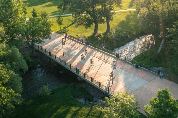 Aerial view of cyclists on a bridge over a stream, surrounded by lush trees and green fields. Warm sunlight casts long shadows, creating a serene atmosphere.
