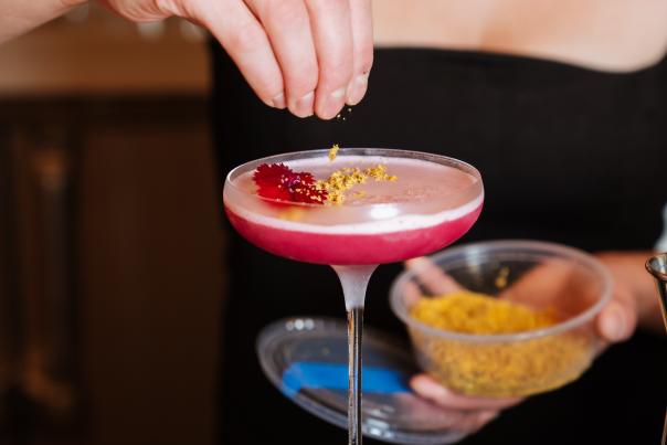 A mixologist tops a bright pink cocktail off with a pinch of yellow bee pollen