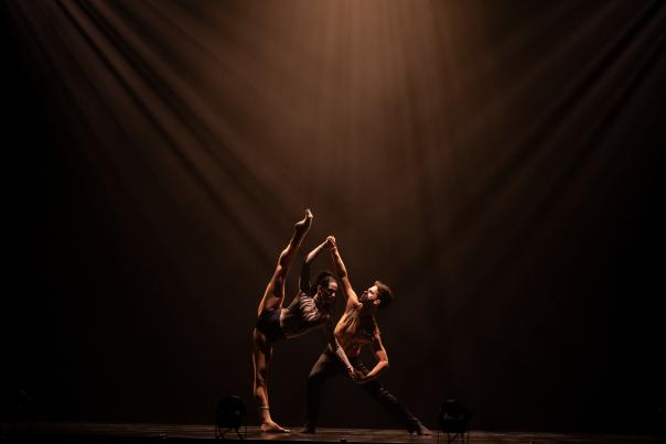Two contemporary dancers perform an expressive lift under dramatic stage lighting.
