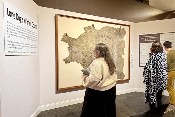 A gallery space with three people standing reading information and inspecting a painting on a hide, framed on the wall.