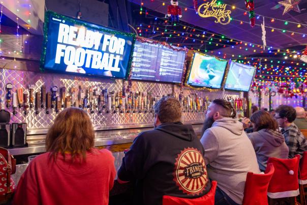 Four patrons sit at the bar at Barley's Kitchen + Tap and look forward at televisions. The bar is decorated with holiday lights and decorations.