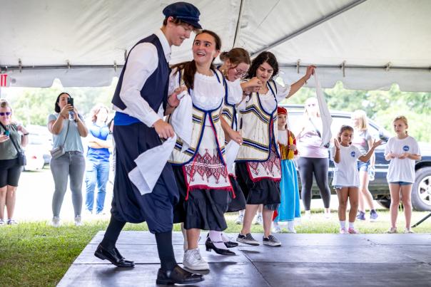 A group of teens in traditional Greek attire perform a lively folk dance under a large festival tent at the Fayetteville Greek Festival, while children and spectators watch and record the moment with their phones in the background.