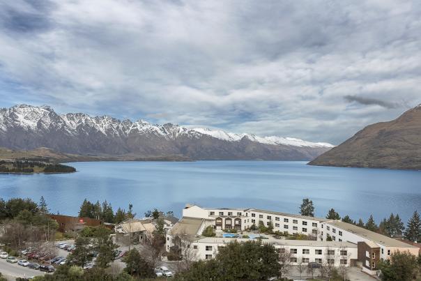A stunning view of Lake Wakatipu with the Remarkables mountain range in the background, featuring a lakeside hotel and surrounding greenery in the foreground.