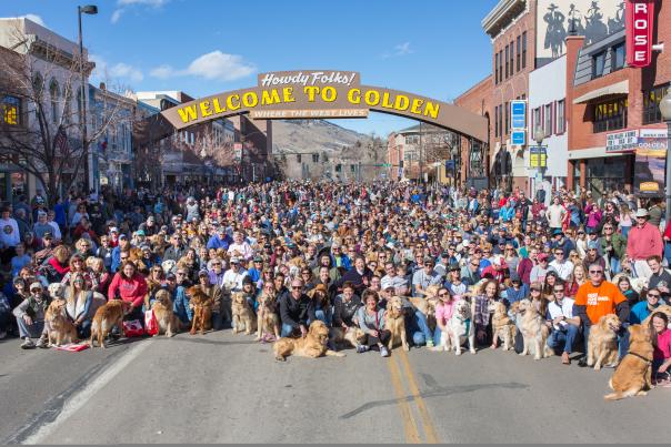 Golden retrievers and owners gather under "Welcome to Golden" Arch