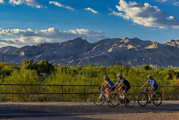 Profile view of three bikers riding on the loop. Greenery, mountains and bright blue sky in the background