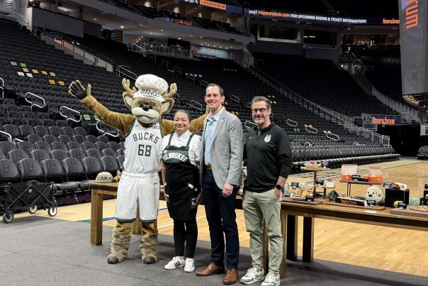 Milwaukee Bucks mascot Bango poses with three people beside a display of food offerings on the basketball court at Fiserv Forum.