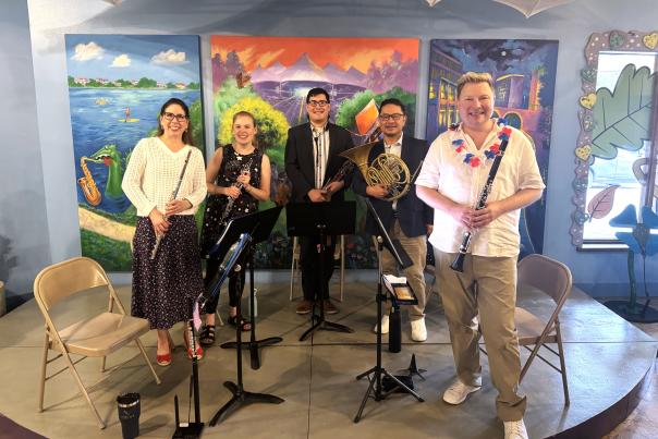 The woodwinds from The Woodlands Symphony smile for the camera after performing at The Woodlands Children's Museum. Five musicians stand on the stage: a woman holding a flute, a woman holding a clarinet, then three men holding a bassoon, French horn, and clarinet.