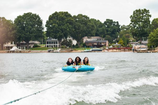 Three people wearing life jackets while tubing on Lake Norman, enjoying a family-friendly watersports adventure with scenic waterfront homes and trees in the background.