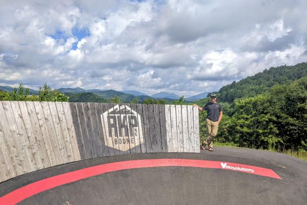 A man stands by a tall wooden wall-ride element at Rocky Knob Mountain Bike Park with a mountain scene in the background.