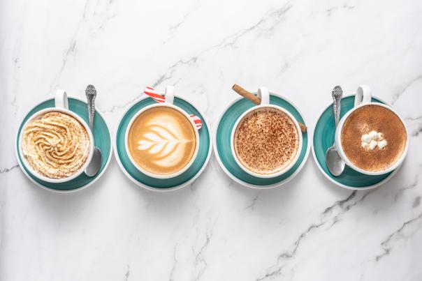 Four lattes are lined up across a white marble table: gingerbread, peppermint, tiramisu, and pumpkin spice.