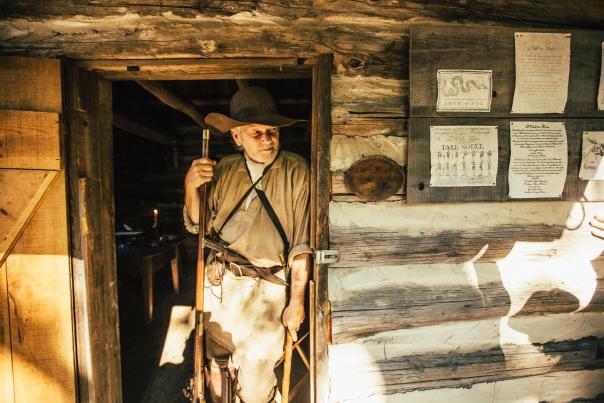 A man leans against the door frame of an old cabin with a long rifle in one hand and a small hatchet lowered in the other hand. He's dressed in frontier garb with a wide brim hat.