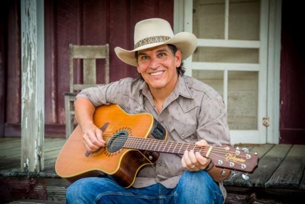 A man in a western shirt and light cowboy hat smiles at the camera as he strums an acoustic guitar from the porch of a rustic red-and-white house.