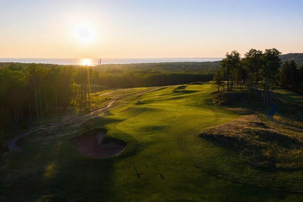 A serene golf course at sunset, with the sun setting over a distant forest and Lake Superior. The scene is peaceful, with lush green fairways and sand bunkers.