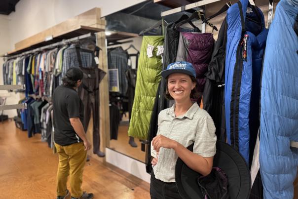 Sturdy used gear owner Kelsey Miller smiles at the camera while a customer looks through outdoor gear on the shelves.