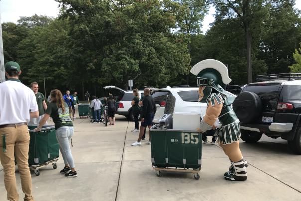 Sparty, the MSU mascot, pushes a move-in bin filled with student belongings while cars with open trunks are parked in the background.