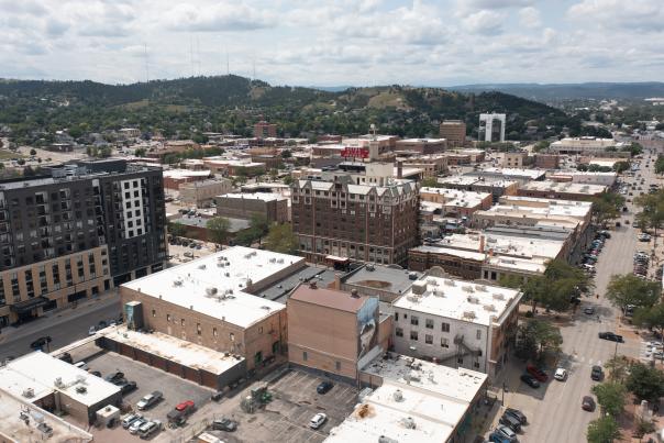An aerial shot of downtown Rapid City, with the Block 5 building and the Hotel Alex Johnson prominently in the foreground, Skyline Drive slightly out of focus, and the silhouette of the Black Hills on the horizon.