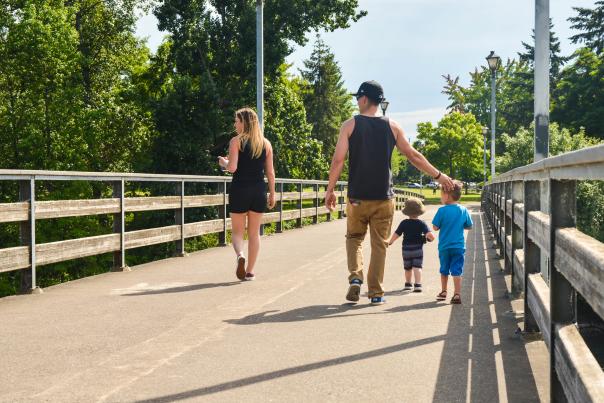 A woman, man and two small children in warm weather clothing walk across a paved pedestrian bridge with wood rails in a park.