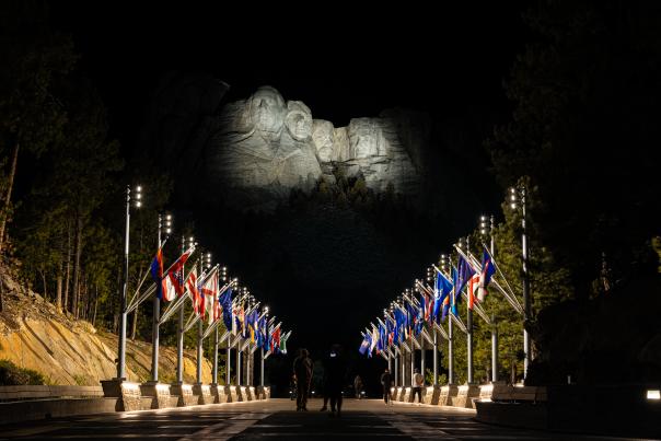 Mount Rushmore illuminated at night, with pathway lined by multiple national flags on both sides. Silhouettes of people walk towards the monument.