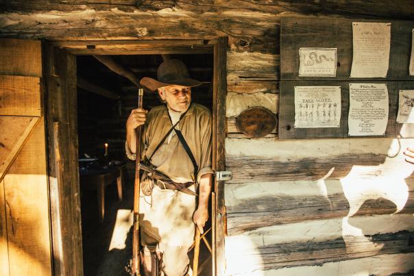 A man leans against the door frame of an old cabin with a long rifle in one hand and a small hatchet lowered in the other hand. He's dressed in frontier garb with a wide brim hat.