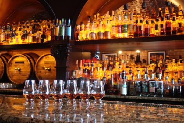 Six tasters of whiskey in front of a wall filled with whiskey barrels and bottles