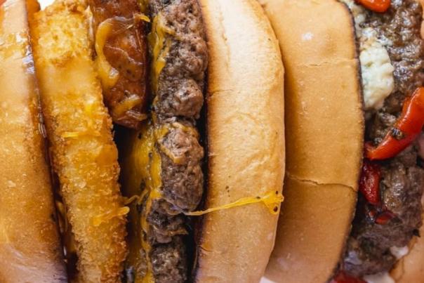 A macro view of the layers of two burgers lined up vertically beside each other like dominoes. There are several layers of buns, beef patties, and cheese as well as a layer of onion rings and grilled peppers.