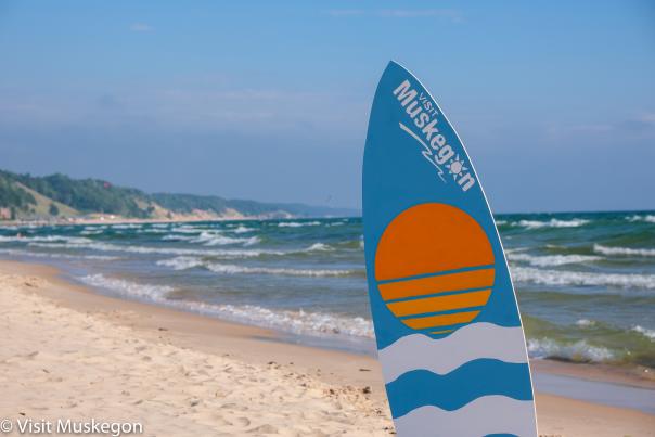 A surfboard with "Visit Muskegon" is planted in the sandy beach. Waves gently crash along the shore, with a backdrop of green hills and a clear blue sky.