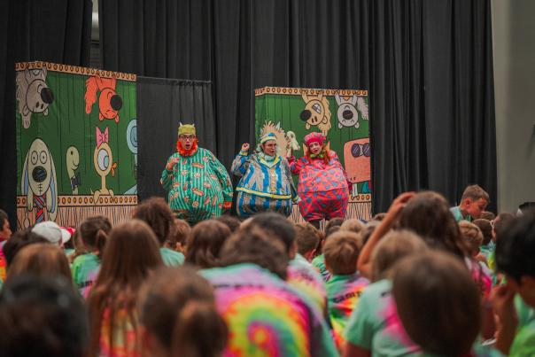 Three actors perform for children at The Pavilion. The actors are singing and dancing, wearing fat suits dressed in pajamas in wacky colors and patterns. The backdrop depicts cartoon animals. Kids in the audience are wearing matching tie-dye shirts.