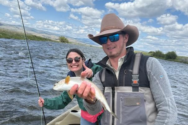 A man in a wide-brimmed hat holds a fish while a woman smiles beside him in a boat on a lake, surrounded by water and clouds.
