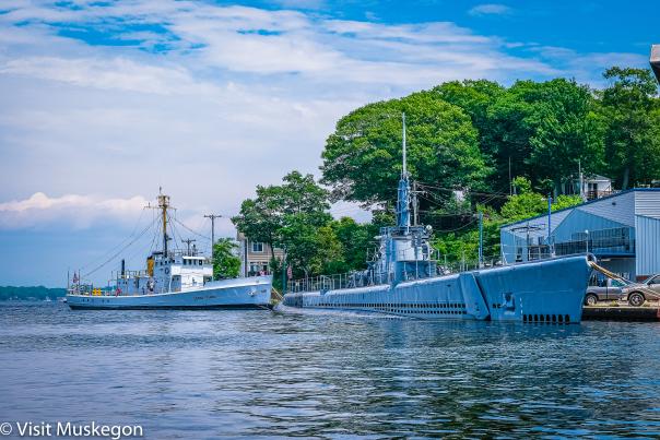 Image of two ships moored at a dock in Muskegon, with one being a historical naval submarine. Clear skies and calm water with buildings in the background.