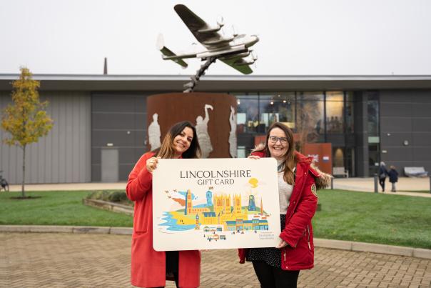 Two people holding a large Lincolnshire Gift Card in front of the International Bomber Command Centre