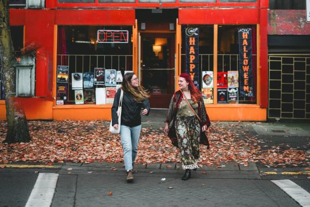 Two people walking past a red storefront with autumn leaves on the ground.
