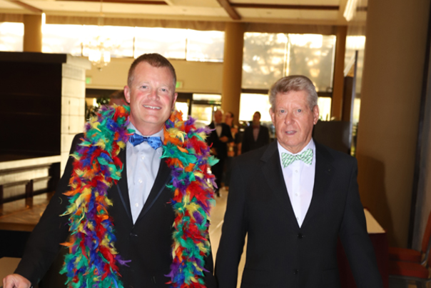 Two smiling gala attendees in tuxedos with rainbow feather boas