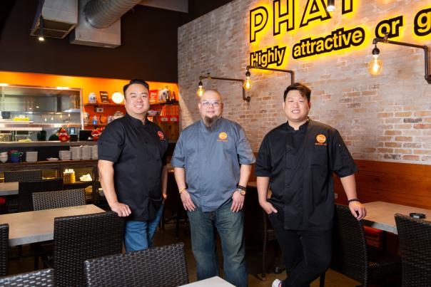 The camera faces Marvin He, Terry Wong, and Kevin Lee under the neon yellow "Phat (adjective) Highly attractive or gratifying" sign inside Phat Eatery.