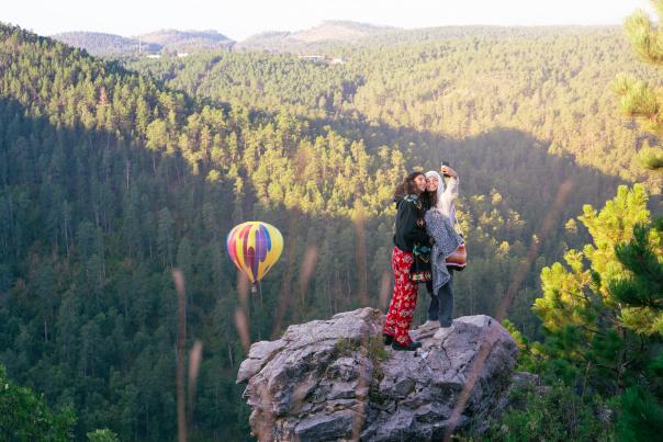 Two spectators taking a selfie as a hot air balloon rises in the black hills behind them