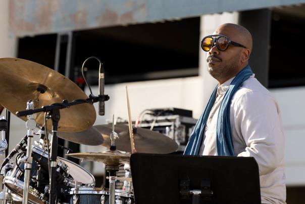 A close up view of a man in sun glasses and a white t-shirt with a blue scarf playing the drums on an outdoor stage.
