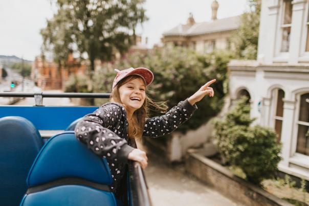 A young girl sat on an open top tour bus pointing and smiling with residential buildings in the background