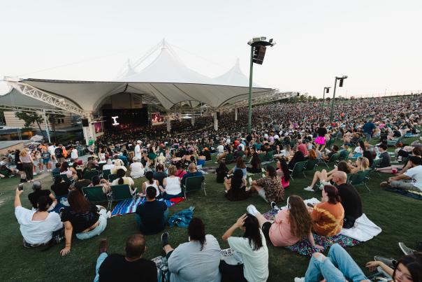 The camera faces The Pavilion stage from the lawn. Both the stands and the lawn are packed with Post Malone fans as they await the start of the concert.