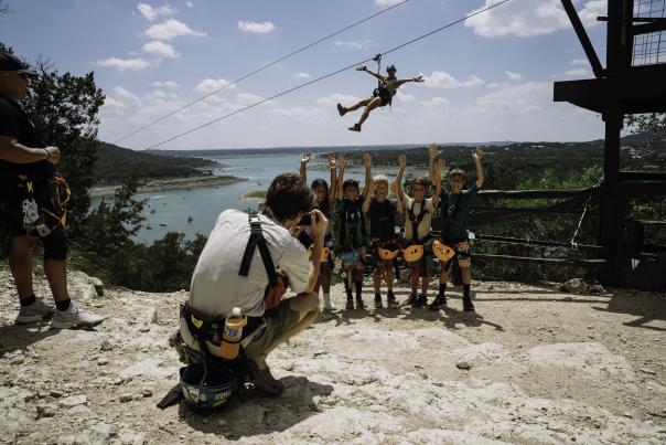 Image of a man taking a group photo of kids at Lake Travis Zipline with someone zipping down behind them.