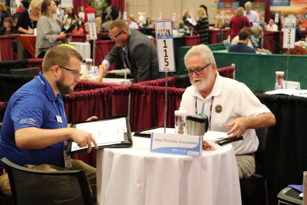 Two men sit at a table in a large meeting space, engaged in a work-related discussion. A sign displaying booth number 115 is on the table.