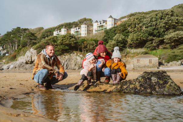 A man, woman and two small girls in winter clothing looking at a rockpool on Caswell beach