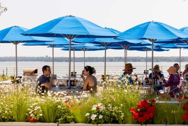 People sitting at an outdoor restaurant that over looks the lake and is surrounding by flowers and greenery.