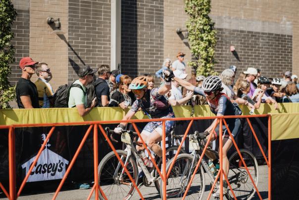 Two cyclists cross a finish line, high-fiving supporters behind a barrier. A crowd watches, with greenery and Downtown Bentonville buildings in the background