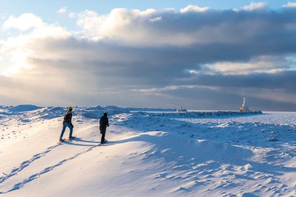 Two people snowshoe frozen Lake Superior shoreline at McLain State Park. The lighthouse can be seen in the background.