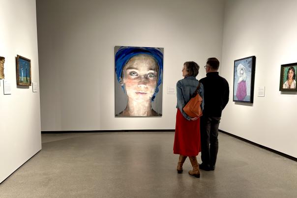 Two visitors at Muskegon Museum of Art viewing various portraits, including a prominent portrait  painting of a young woman with a blue scarf covering her head.