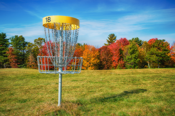 18th hole disc golf cage with yellow top sits in grassy field with fall trees in background