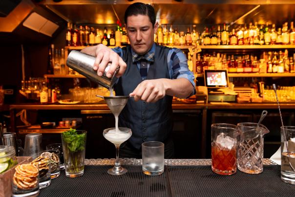 Bartender pours a light drink from a shaker through a filter into a martini glass on a bar counter. Other cocktail glasses and garnishments can also be seen on the counter. In the background, there are two shelves stacked full of liquor bottles.